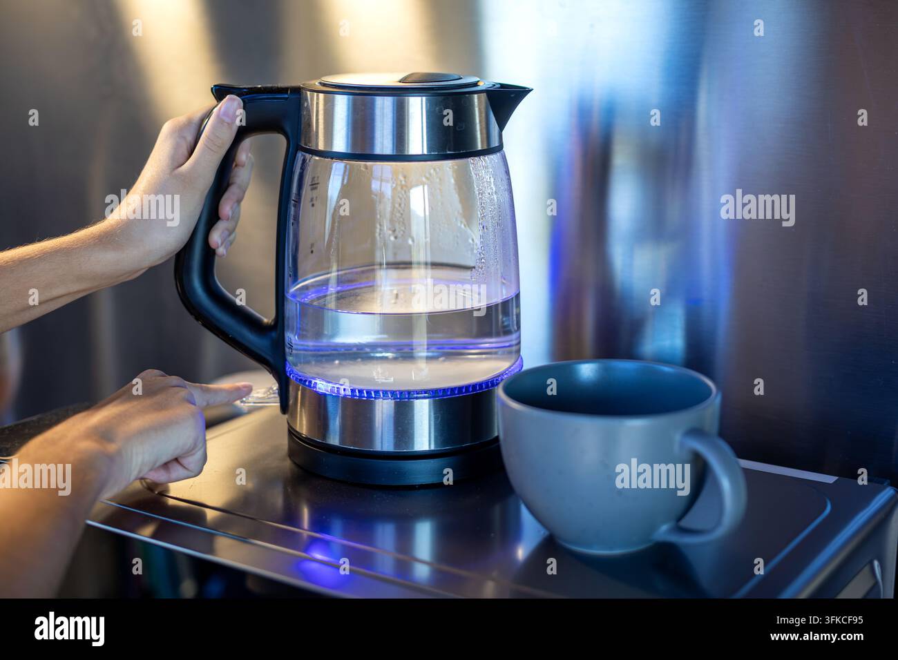 Person utilisant une bouilloire électrique en verre à côté d'une tasse en céramique. Personne tenant une bouilloire électrique en verre remplie d'eau tout en appuyant sur un bouton de contrôle à côté Banque D'Images