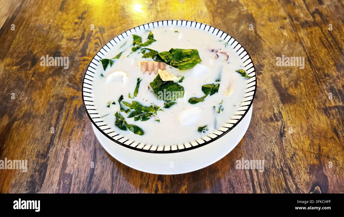 Feuilles de bai liang thaïlandaises avec des calmars cuits dans du lait de coco crémeux, servis dans un bol en céramique sur une table en bois, un plat traditionnel du sud de la Thaïlande - Image de stock capturée avec un smartphone