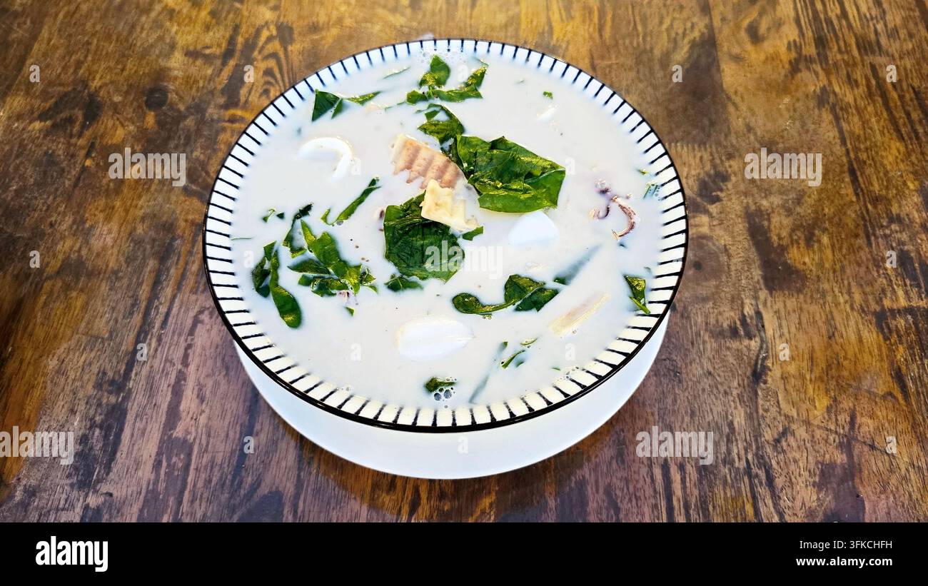 Feuilles de bai liang thaïlandaises avec des calmars cuits dans du lait de coco crémeux, servis dans un bol en céramique sur une table en bois, un plat traditionnel du sud de la Thaïlande - Image de stock capturée avec un smartphone