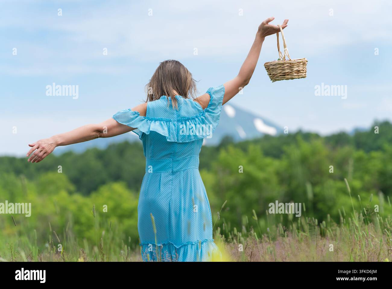 Vue arrière d'une femme en robe à pois bleus profitant de l'été en plein air, les bras écartés tenant un panier en osier. Bras féminins grands ouverts dans une pose joyeuse Banque D'Images
