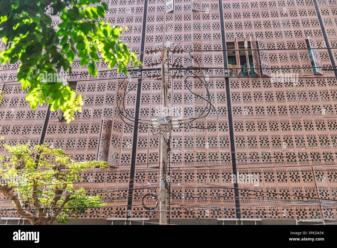 Une façade de bâtiment en briques à motifs à Bangkok est traversée par des câbles utilitaires denses et partiellement encadrée par un feuillage baigné de soleil. Banque D'Images