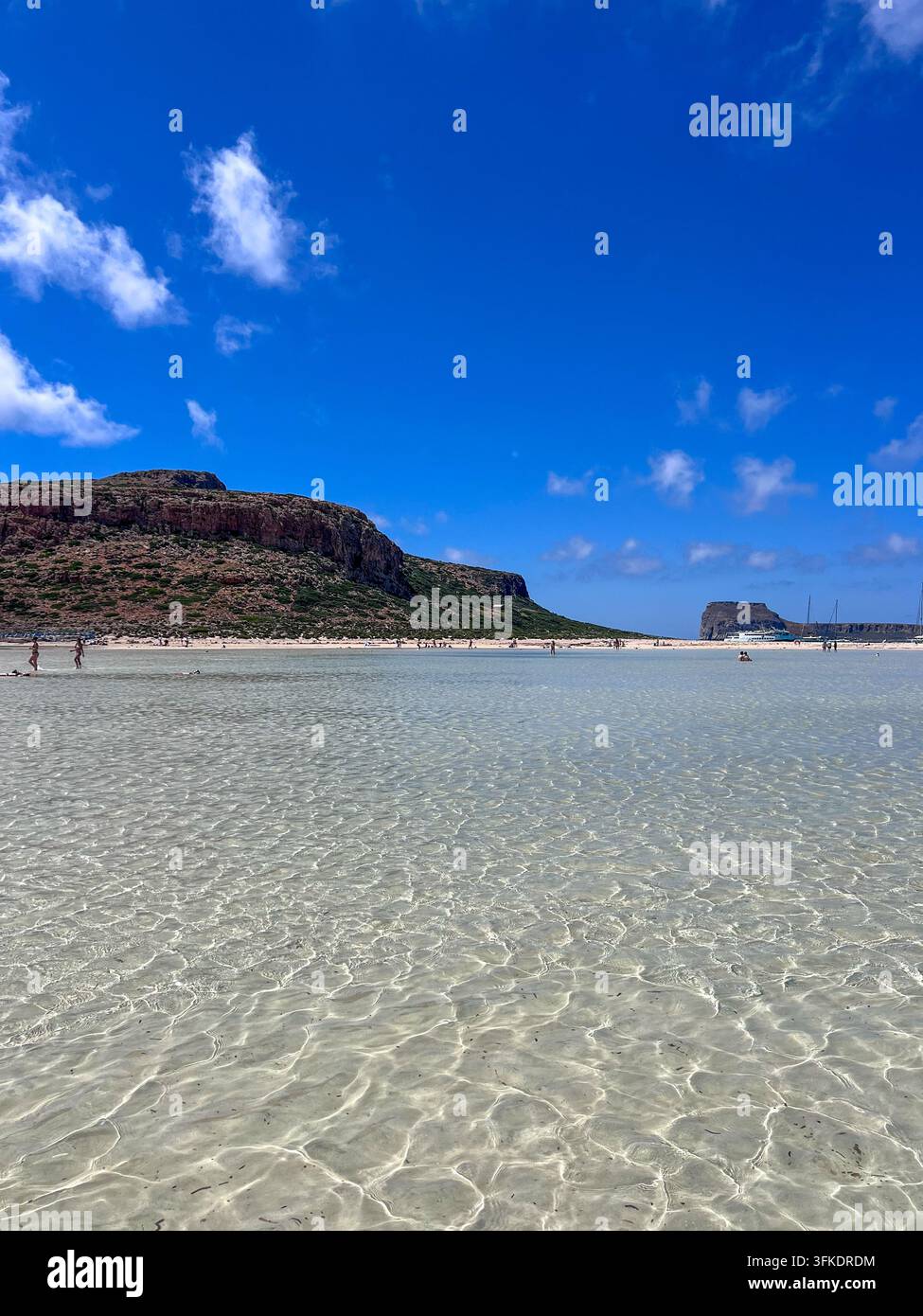 Des gens pataugeant dans une eau claire comme du cristal peu profonde à Balos Beach, Crète, avec une rive sablonneuse et une pointe rocheuse en arrière-plan. - Image de stock capturée avec un smartphone