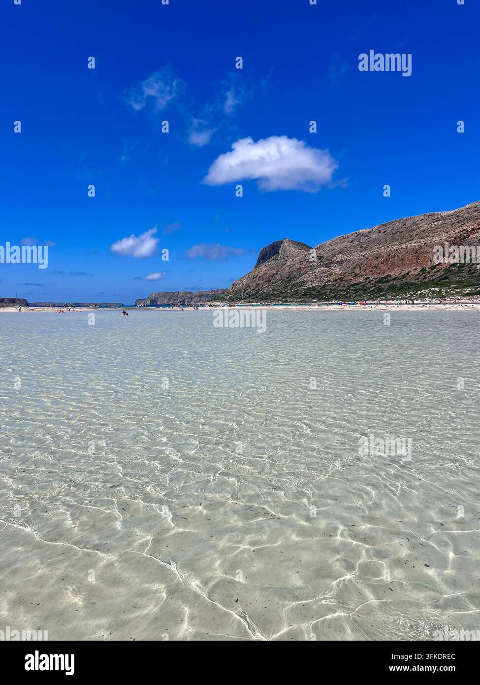 Eau peu profonde et cristalline à Balos Beach, Crète, avec un fond sablonneux visible, des baigneurs lointains et des montagnes accidentées. ( - Image de stock capturée avec un smartphone