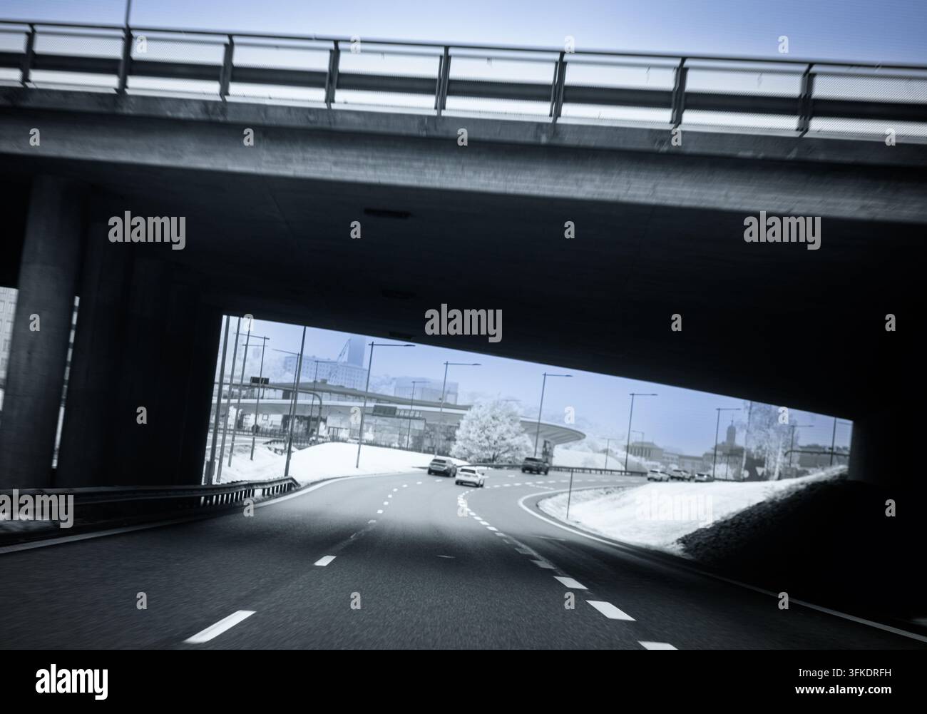 Une scène monochrome proche de l'infrarouge montre des voitures roulant le long d'une route sous une passerelle en béton. La neige couvre le sol à l'arrière-plan avec des arbres visibles Banque D'Images