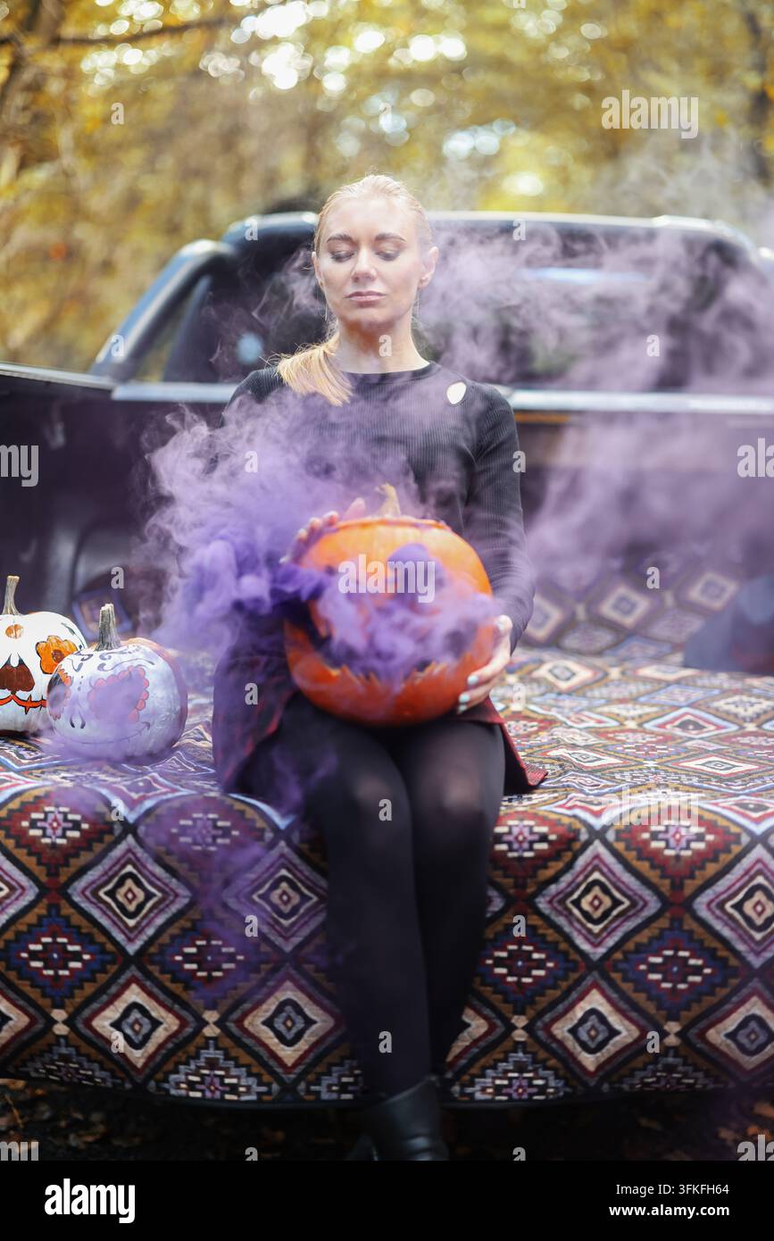 Femme élégante avec un potiron sculpté effrayant et de la fumée violette, idéale pour la publicité Halloween, les événements et les promotions saisonnières. Banque D'Images