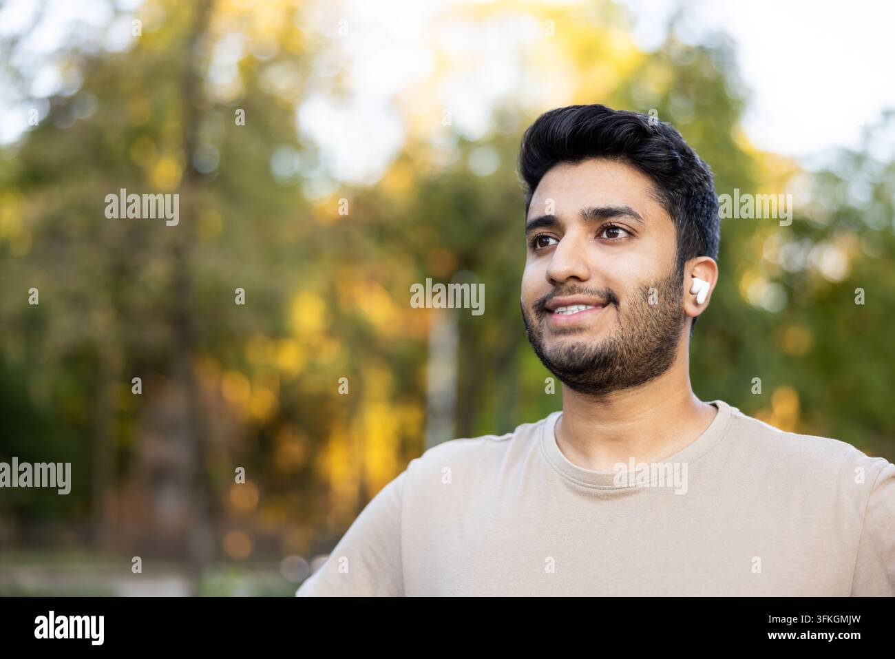 Jeune homme souriant portant des écouteurs sans fil profitant de contenus audio en se détendant dans un parc, représentant le style de vie, la technologie et la connexion avec la nature Banque D'Images