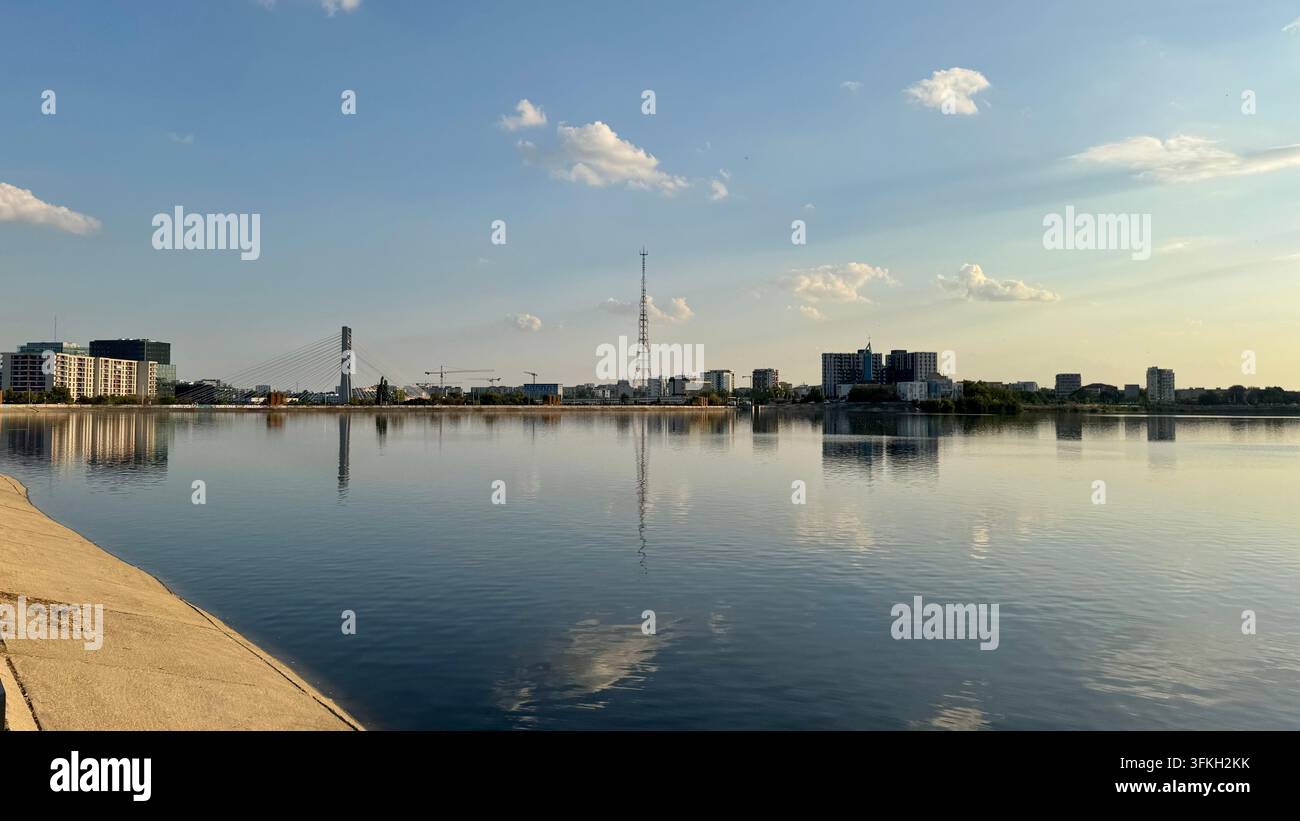 Le lac Morii (Morii Lake) reflète un ciel bleu et des nuages blancs, avec l’horizon de Bucarest visible à travers l’eau. Banque D'Images