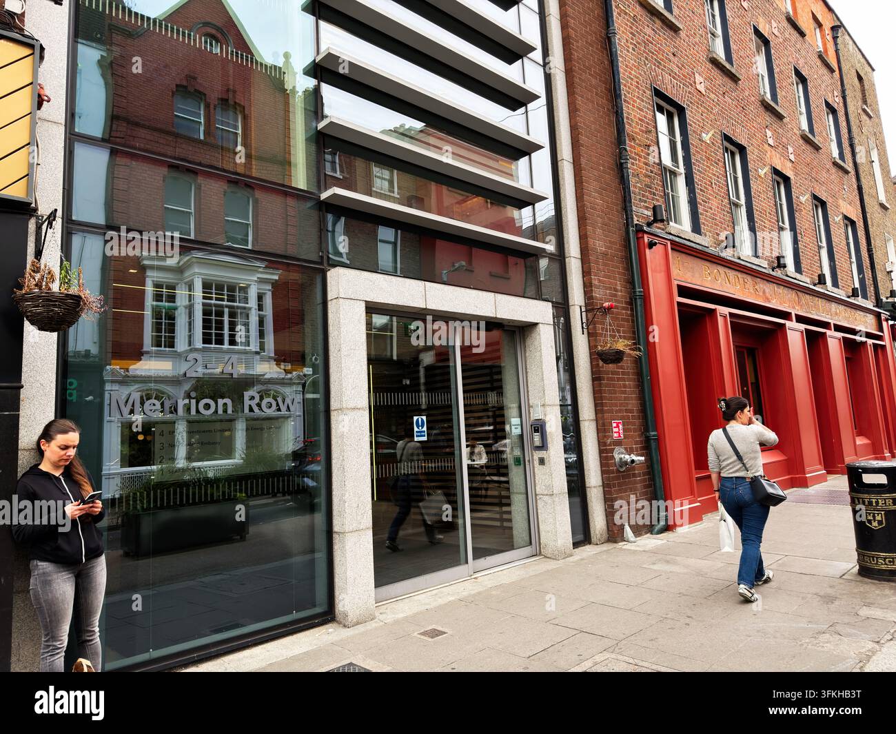 Dublin, Irlande - 22 mai 2026 : Des piétons marchent et vérifient leurs téléphones devant une entrée de bureau en verre moderne de Merrion Row à côté d'une façade rouge historique à D Banque D'Images