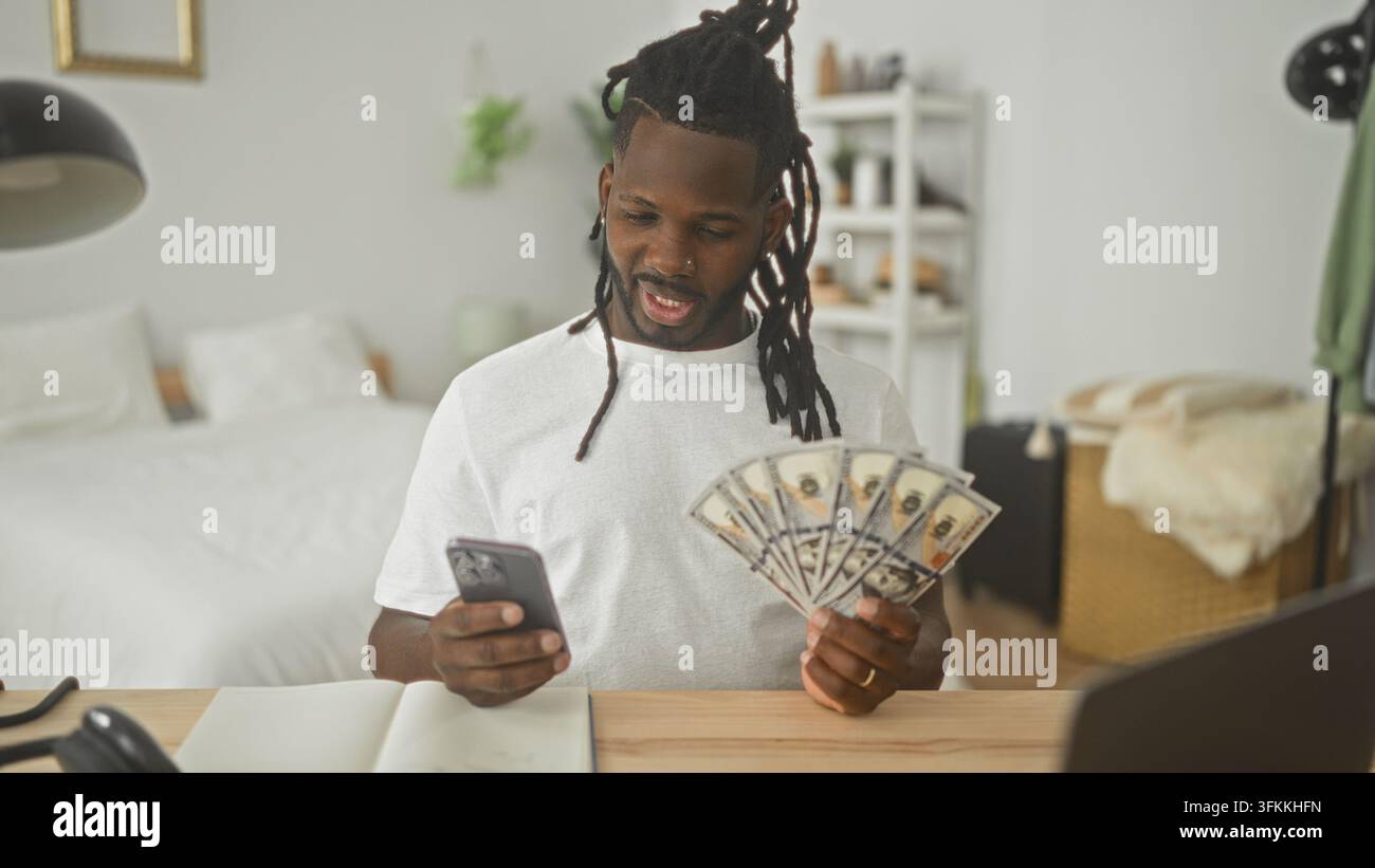 Homme éparpille des dollars tout en vérifiant son smartphone dans une chambre avec un notebook et un ordinateur portable sur le bureau ; célébration de joie et de succès. Banque D'Images