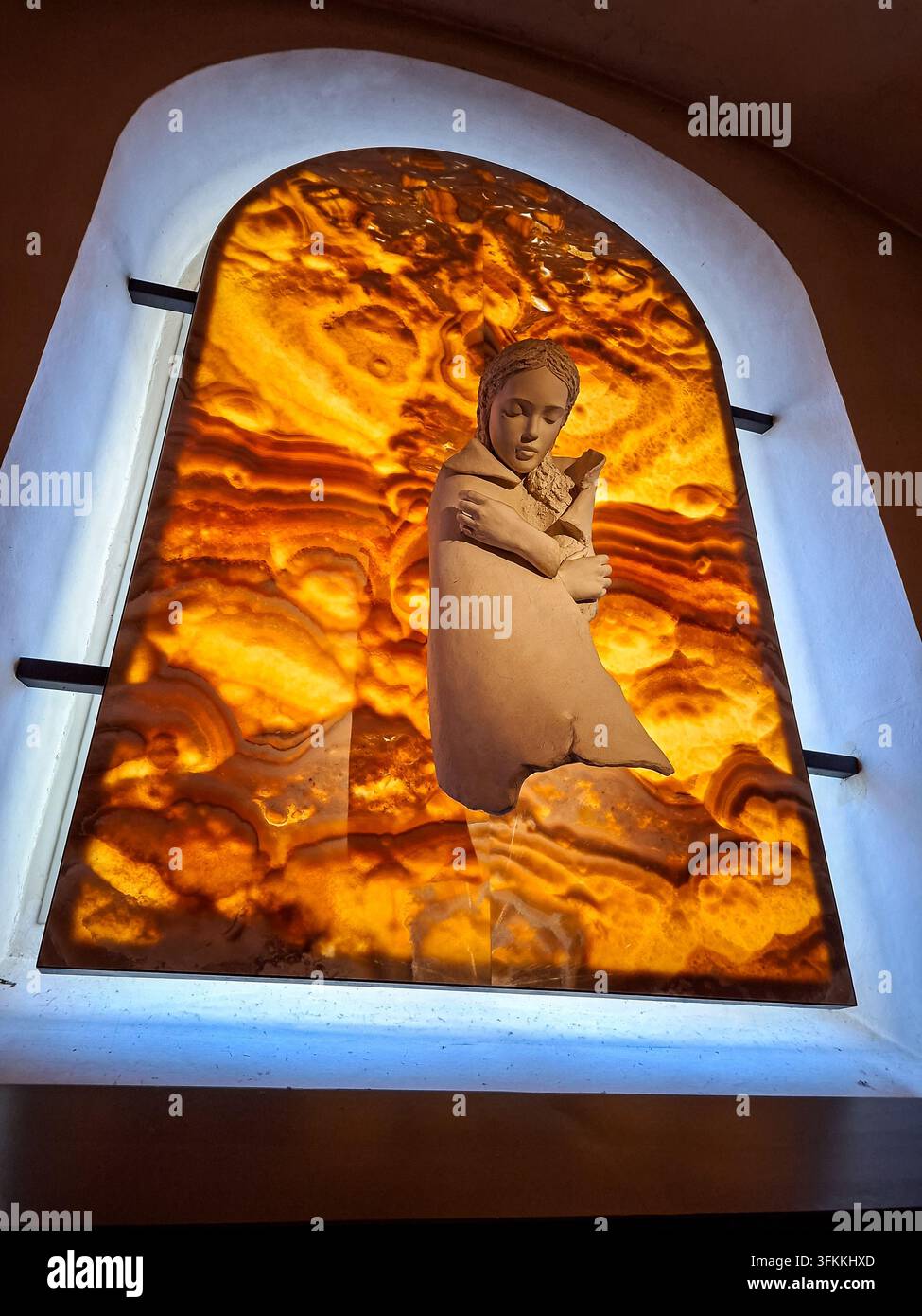 Un plan rapproché d'une figure sculptée d'une femme s'embrassant elle-même, positionnée devant un panneau d'onyx vibrant et illuminé dans une fenêtre en arc. T Banque D'Images