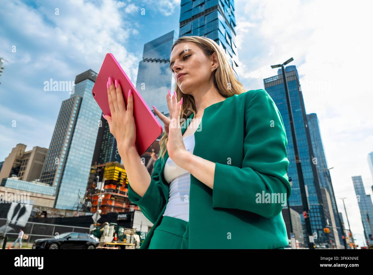 Dirigeante féminine en costume vert se tient à Manhattan avec une tablette rose, gesticulant avec assurance devant des gratte-ciel imposants, symbolisant les affaires modernes Banque D'Images