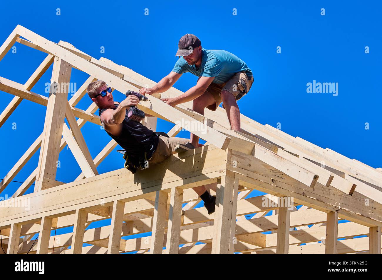 La charpente en bois constitue l'élément structurel principal. Un ouvrier utilise une perceuse sans fil pour fixer les éléments tandis qu'un collègue maintient en place le pied de la poutre. Banque D'Images