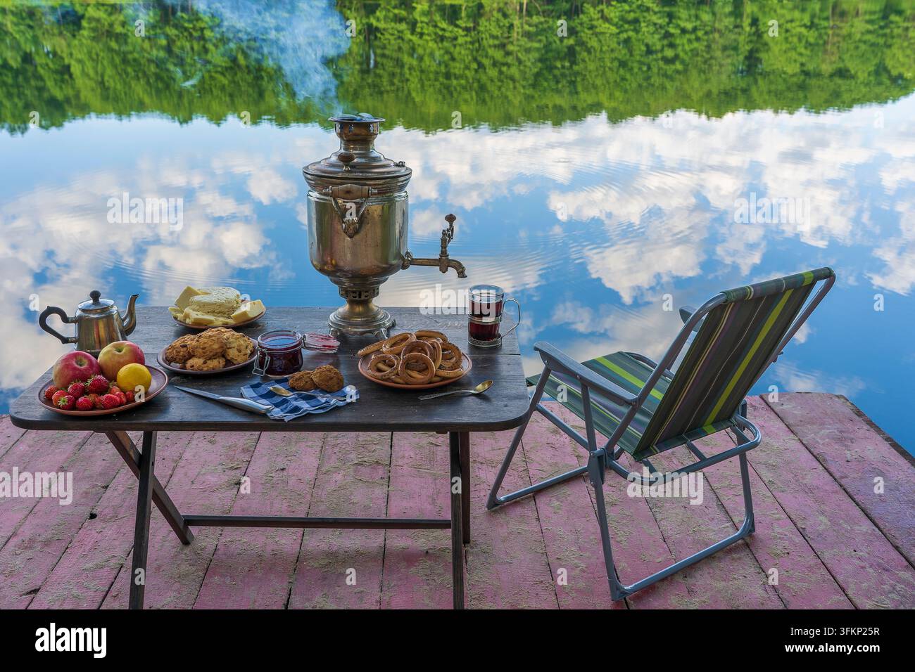 Samovar vintage en métal avec fumée blanche et nourriture sur la table près du lac d'eau calme dans la forêt verte au matin, Ukraine. Concept de nourriture naturelle Banque D'Images