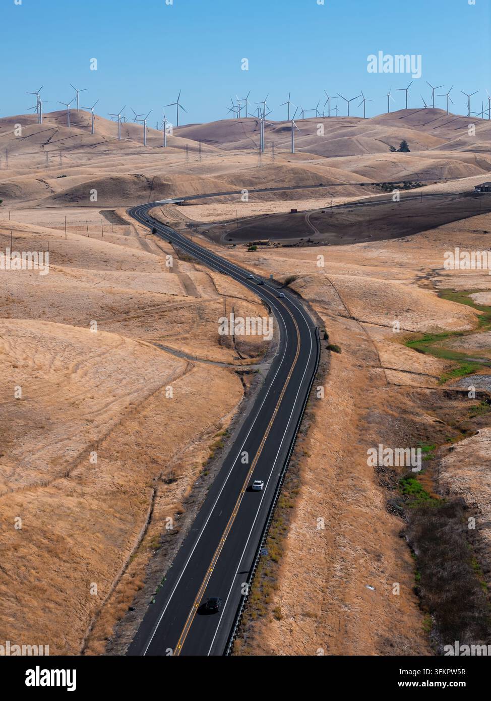 Une vue aérienne montre une autoroute pavée sinuant à travers des collines arides menant vers des éoliennes produisant de l'électricité renouvelable à travers un paysage en Ca Banque D'Images