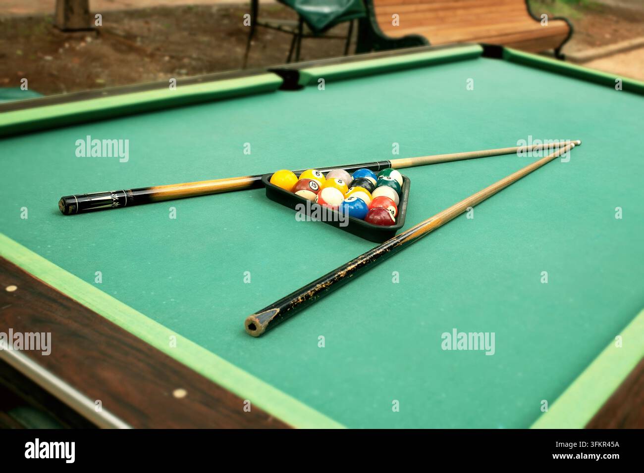 Installation de table de billard d'extérieur avec une toile verte usée et tachée, boules de billard colorées disposées en triangle, mise au point sélective sur les queues dans un parc automnal Banque D'Images