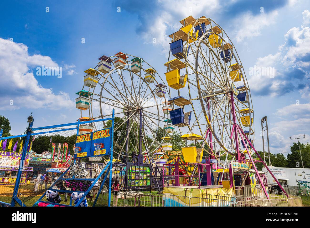 Deux grandes roues de Ferris se dressent de manière proéminente sur un terrain de foire en plein air sous un ciel partiellement nuageux. „BASKETBALL BASKETBALL“ apparaît sur un stand de jeu. Banque D'Images
