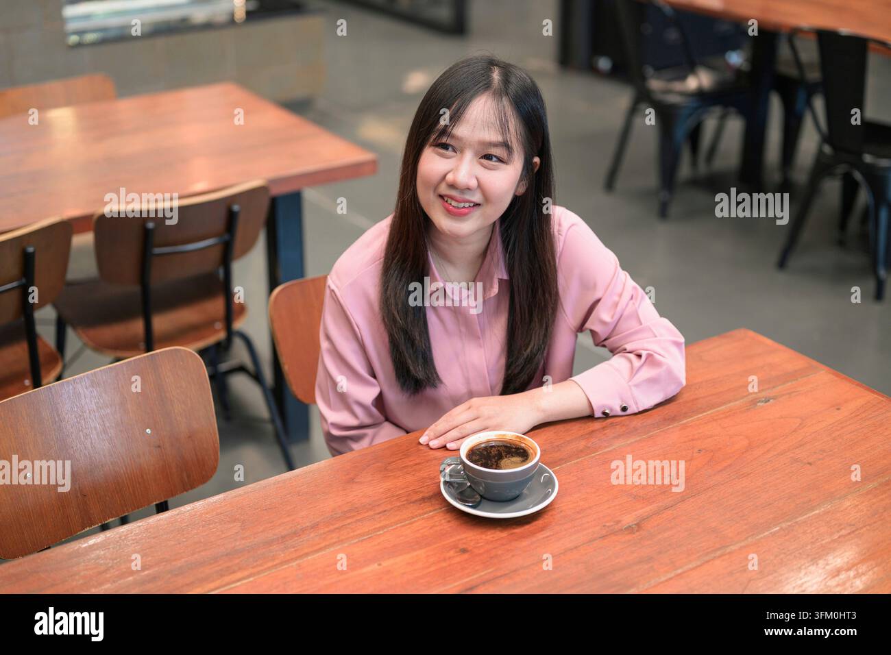 Vu sous un angle élevé, une jeune femme asiatique heureuse en chemise rose est assise à une table en bois dans un café, souriant tout en regardant sur le côté à côté d'une tasse Banque D'Images