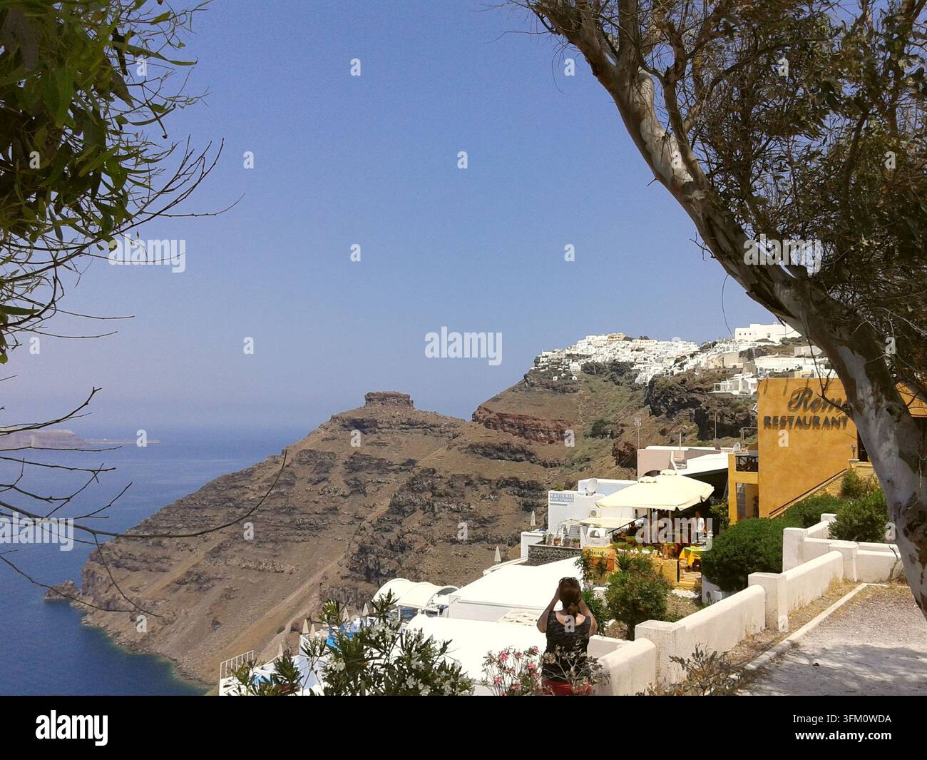 Vue d'un point de vue surplombant la caldeira de Santorin, Grèce, montrant une falaise, des maisons blanches, une enseigne de restaurant et un touriste prenant des photos. Banque D'Images