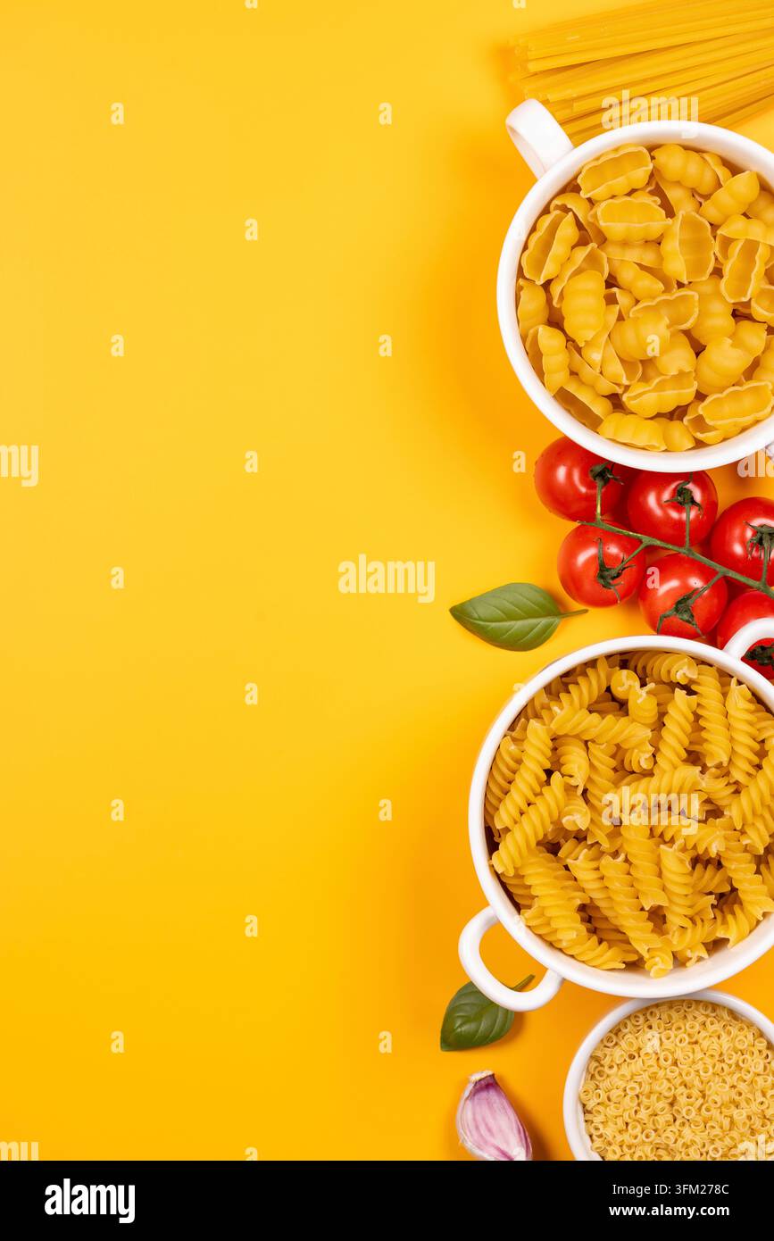 Formes de pâtes crues assorties dans des bols blancs avec tomates fraîches, feuilles de basilic, ail et spaghetti sur un fond jaune vif créant un cul vibrant Banque D'Images