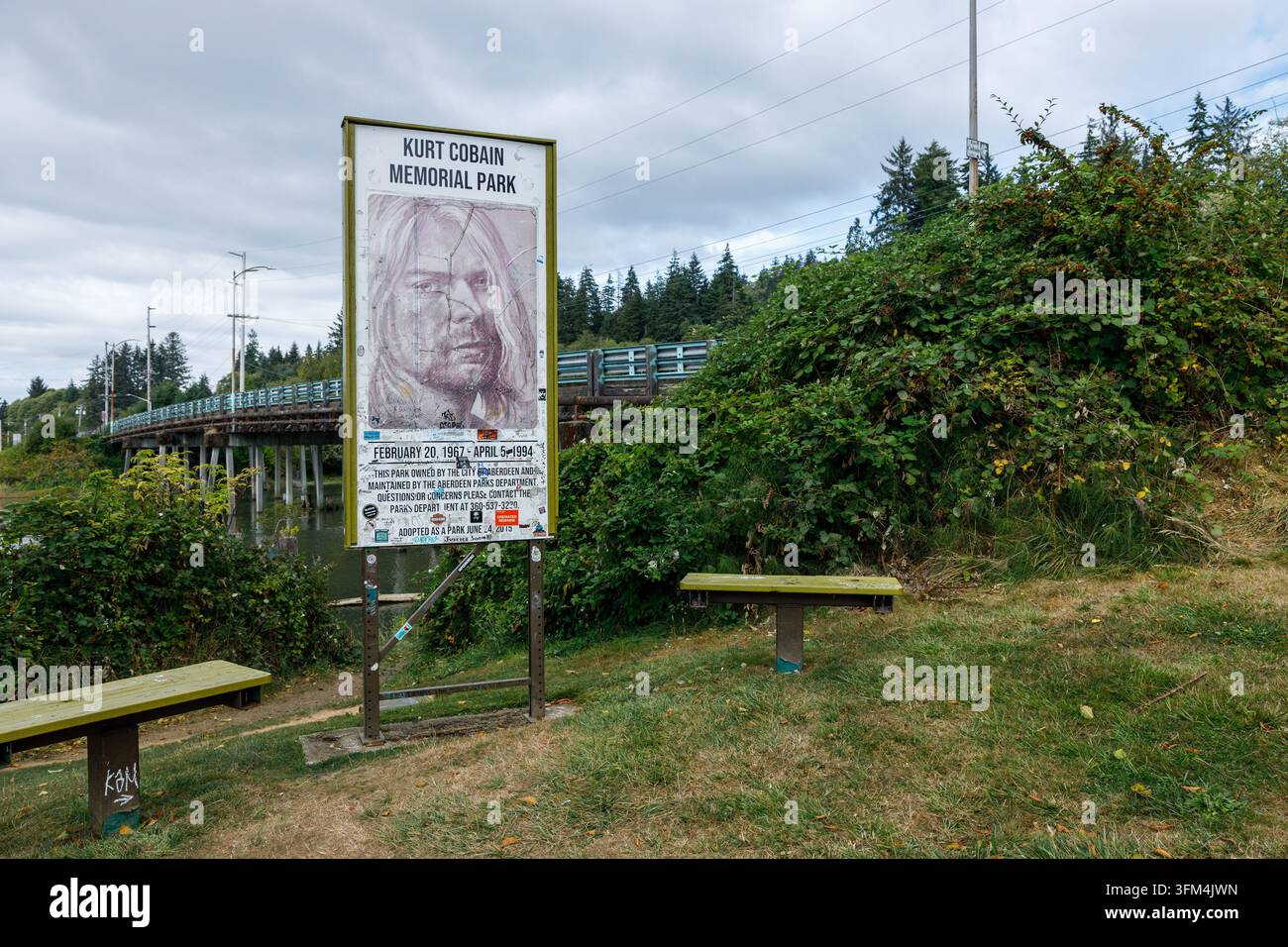 Aberdeen, États-Unis - 30 août 2026 : Vue large du parc commémoratif de Kurt Cobain avec son panneau portrait, deux bancs verts et un pont sur la rivière sous un ciel nuageux Banque D'Images