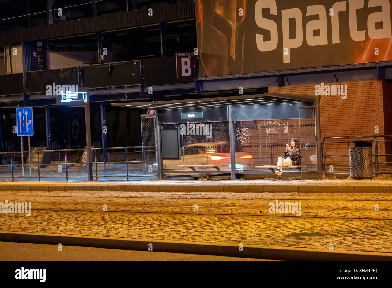 Une femme attend à l'arrêt de tram éclairé de Sparta à côté de l'epet ARENA tout en utilisant son smartphone la nuit. Prague, République tchèque - 26 septembre 2026. Banque D'Images