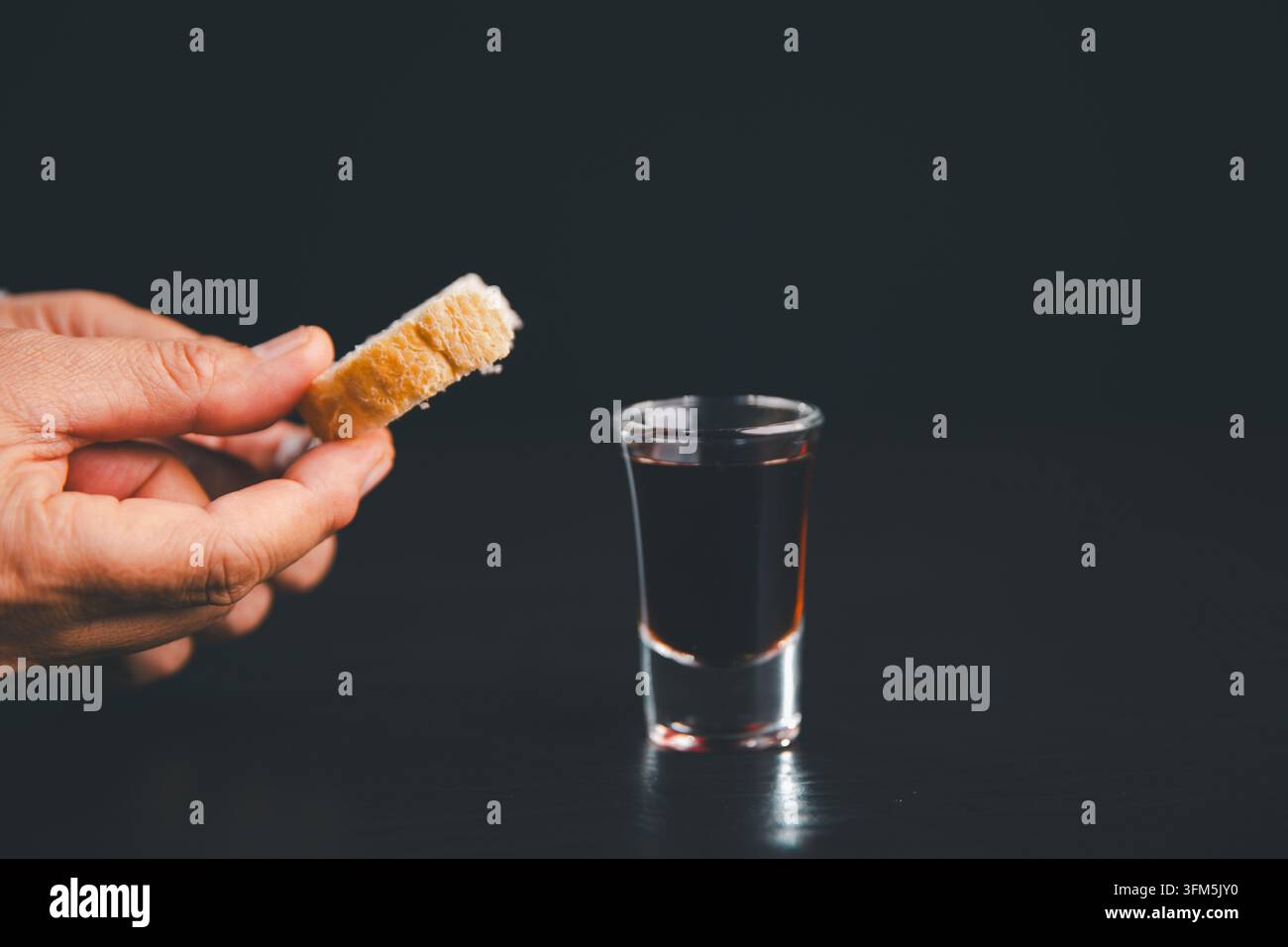 Mains tenant un morceau de pain à côté d'un petit verre de boisson de communion rouge foncé sur un fond noir, illustrant la foi chrétienne, le culte, la prière Banque D'Images