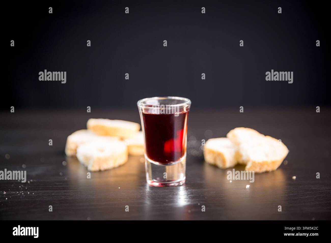 Boisson rouge foncé dans un verre transparent encadré par du pain tranché sur une table en bois noir, créant une nature morte communiale minimale pour la foi chrétienne, sacrée Banque D'Images