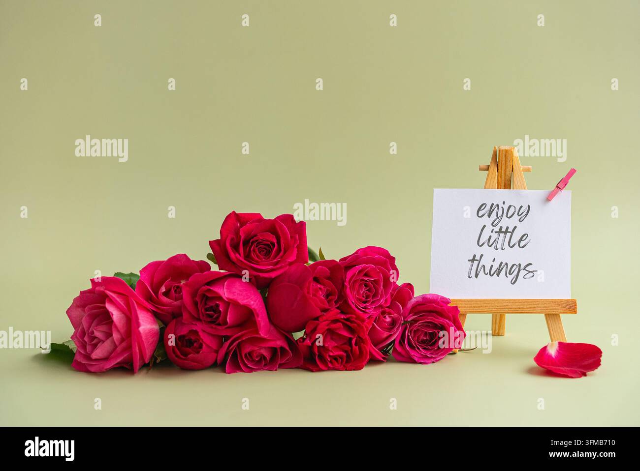 Carte de citation inspirante « ENJOY LITTLE THINGS » affichée sur un chevalet en bois entourée de roses roses vibrantes et de pétales épars sur un fond pastel doux. Composition florale élégante avec message motivant, espace de copie généreux et style minimal adapté aux cartes de vœux, réseaux sociaux, bien-être, soins personnels, encouragement, célébrations, branding et campagnes saisonnières. Banque D'Images