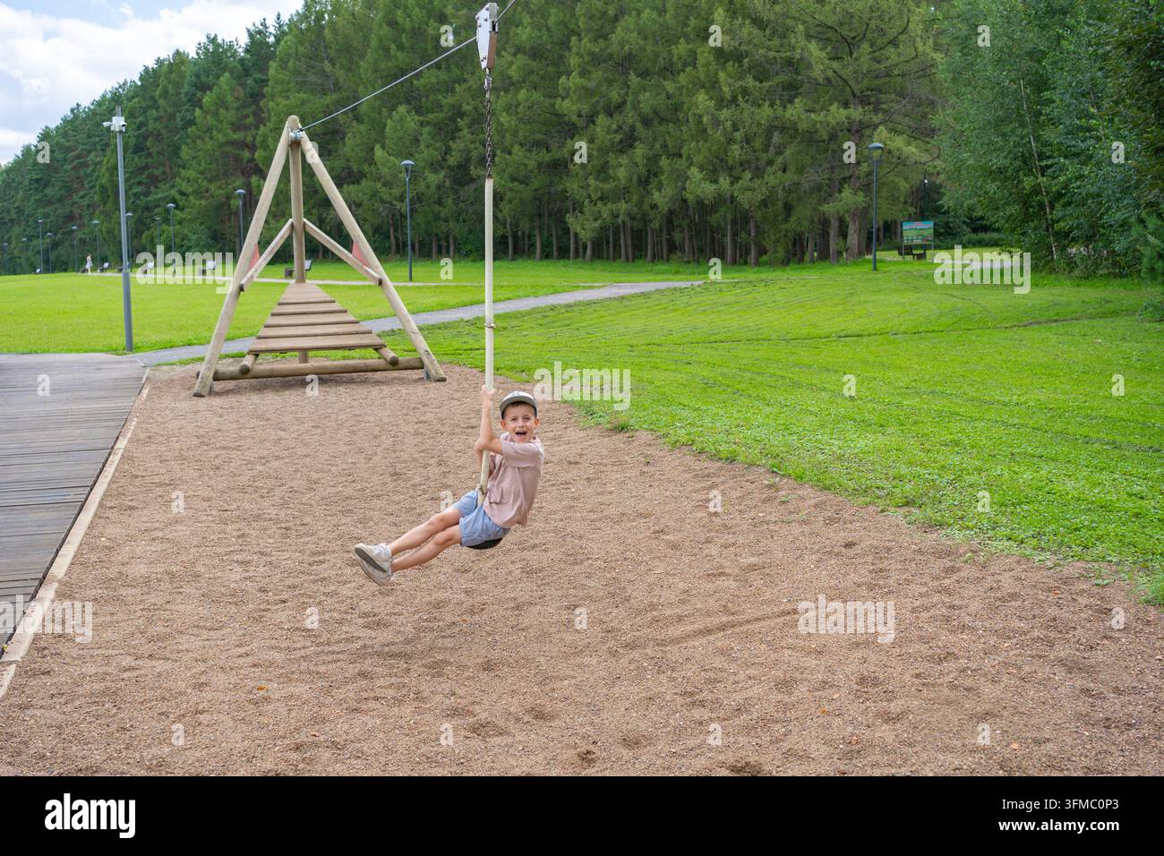 Garçon se balançant, dans le parc, avec une balançoire, plateforme en bois, champ d'herbe, arbres, vu de côté, avec des accents verts, bruns et une ambiance joyeuse Banque D'Images
