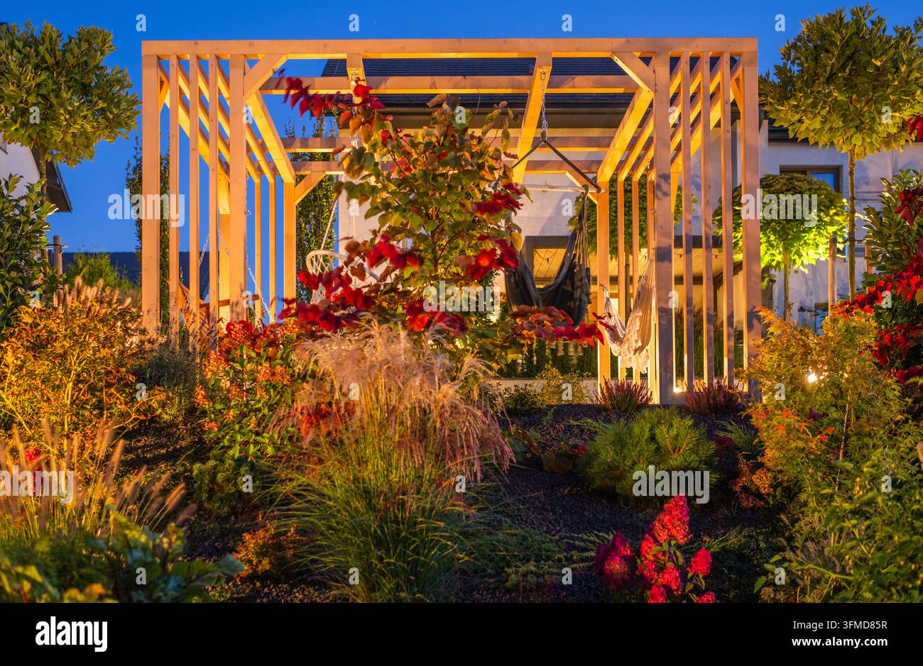 Un éclairage chaud met en valeur une pergola de jardin en bois entourée de plantes grimpantes, de graminées et d'arbustes à l'heure bleue. Banque D'Images