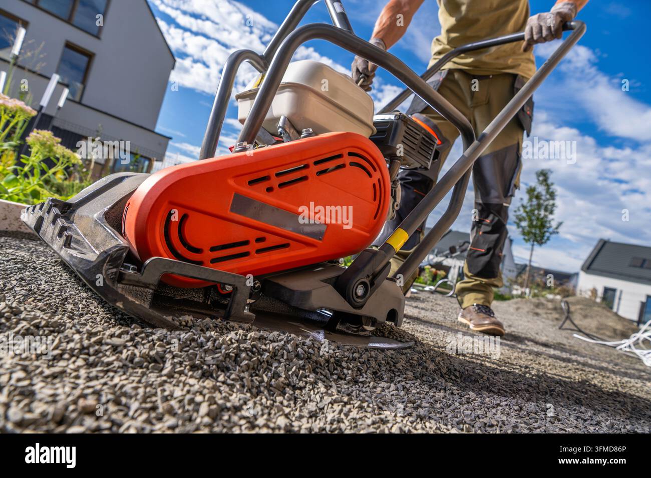 Vue en contre-plongée d'un ouvrier manœuvrant un compacteur à plaque vibrante sur une base de gravier dans un jardin résidentiel. Banque D'Images