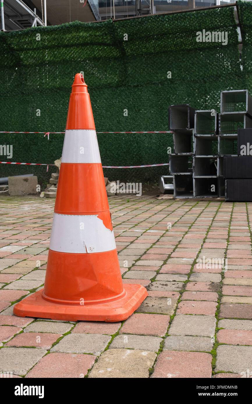 Cône de signalisation orange vif et blanc posé sur un sol pavé près d'une clôture pour avertissement de sécurité. Banque D'Images