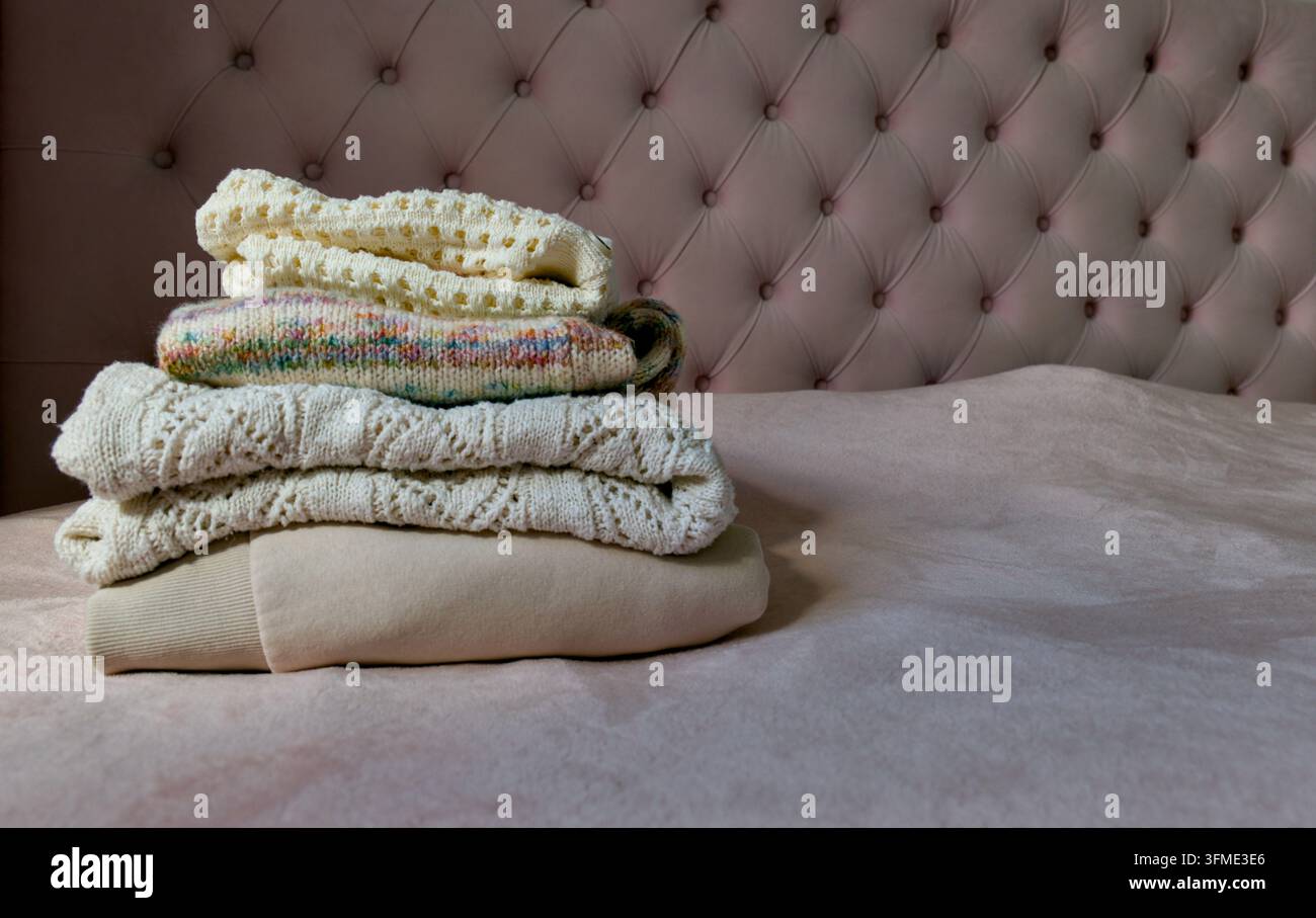 Pile soigneusement empilée de pulls pliés et sweat-shirt beige sur un lit rose confortable avec un fond élégant. Banque D'Images