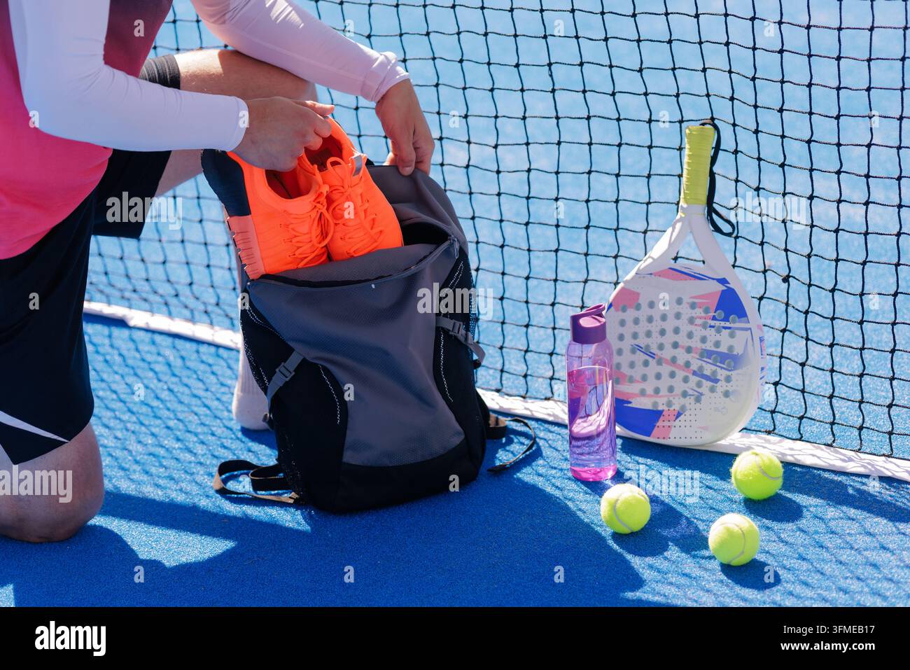 Le joueur de padel se prépare à l'entraînement à côté du filet de court avec des chaussures, une raquette, des balles, un sac à dos et une bouteille d'eau, soutenant les campagnes d'équipement sportif. Banque D'Images