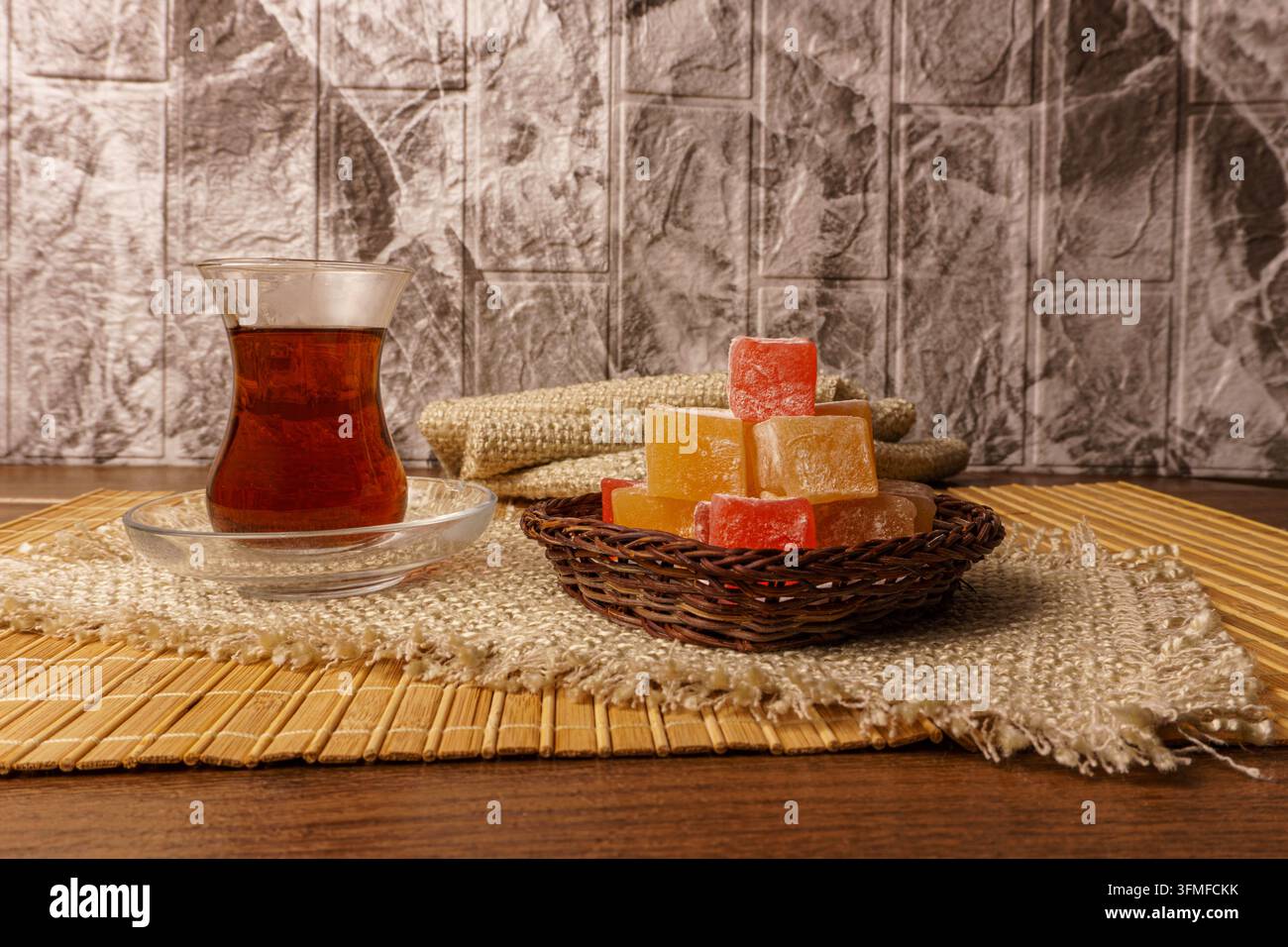 Composition mettant en vedette du thé turc dans une tasse en verre « armudu » et un tas de lokum coloré dans un panier tissé. Servi sur un tapis en bambou et un tissu rugueux agai Banque D'Images