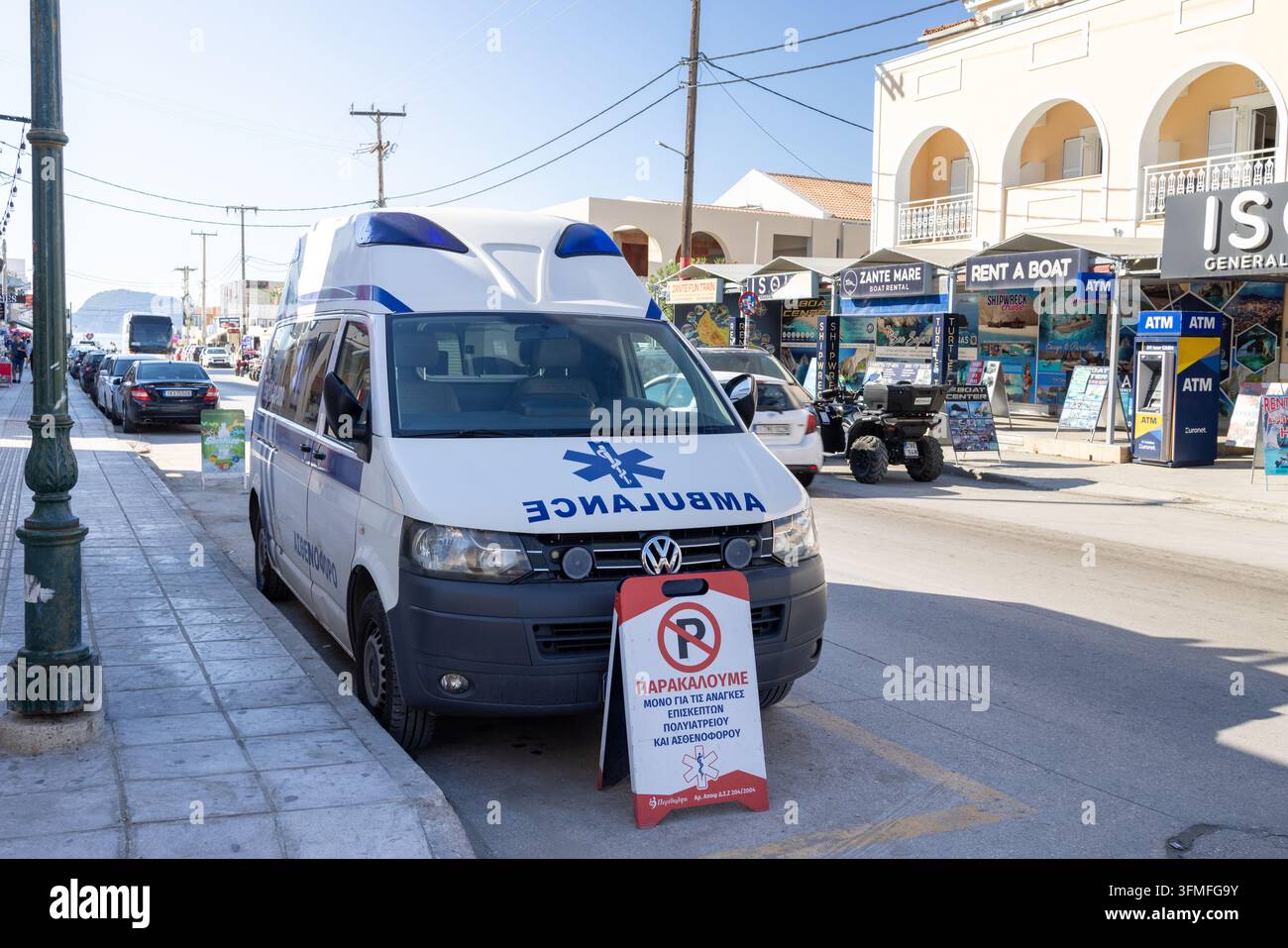 Laganas, Zákynthos Grèce, 1er septembre 2026 : Ambulance garée à côté d'une rue animée de Laganas avec panneaux de location de bateaux, distributeurs de billets, magasins, véhicules, poteaux électriques Banque D'Images