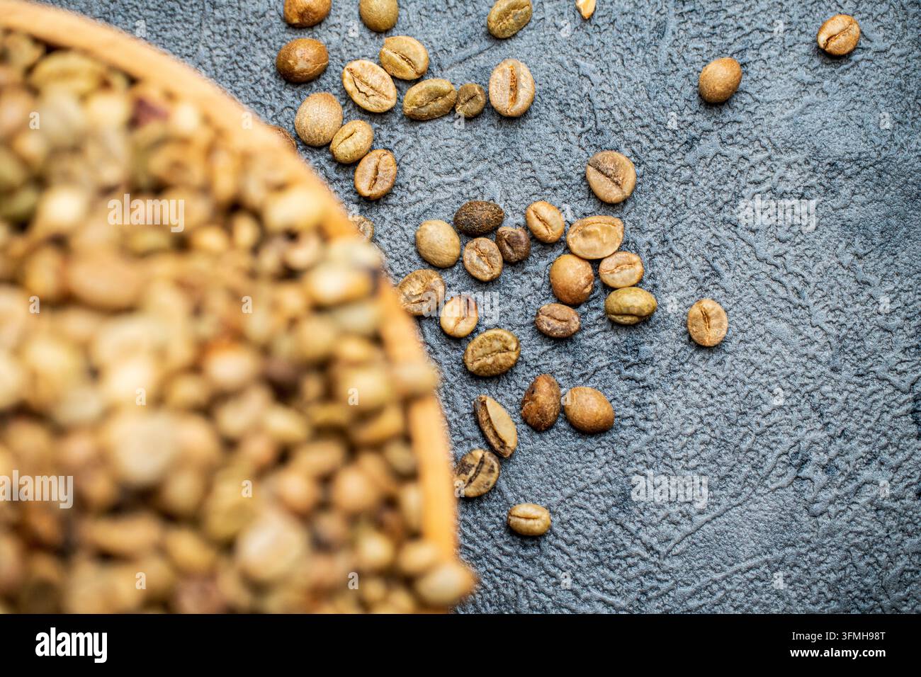 Grains de café verts crus non torréfiés débordant d'un bol en bois sur une surface sombre avec un fond flou de grains éparpillés pour un thème agricole. Banque D'Images