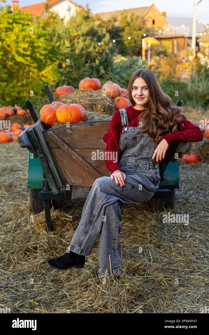 Jeune femme en salopette en jean posant à côté d'une charrette rustique remplie de citrouilles et de foin, portrait chaleureux de récolte automnale à la lumière chaude du soir. Banque D'Images