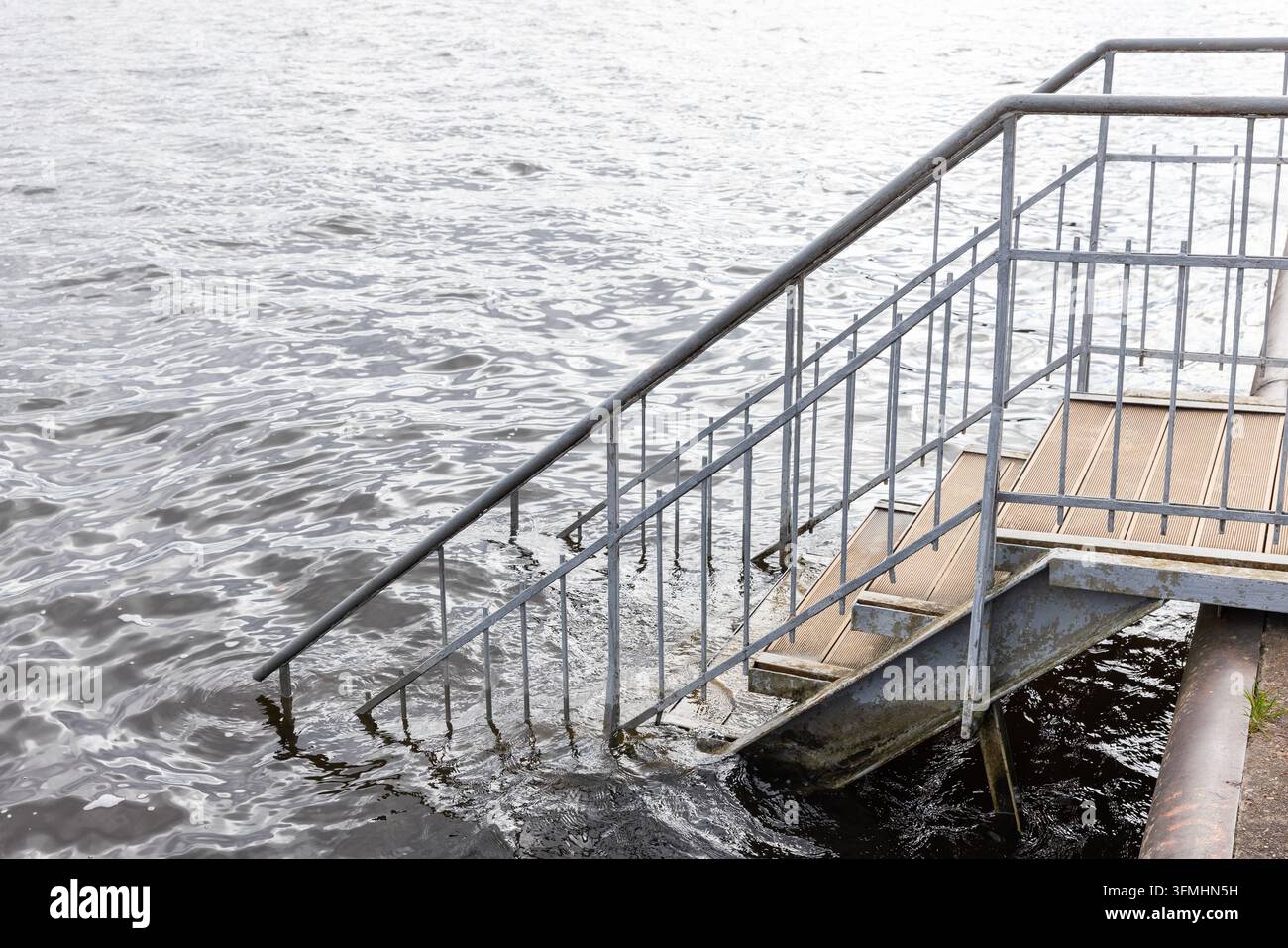 Un escalier en métal rouillé avec des rampes descend d'un quai dans l'eau ondulante de la rivière, partiellement submergé et érodé, évoquant une ambiance sombre et négligée Banque D'Images