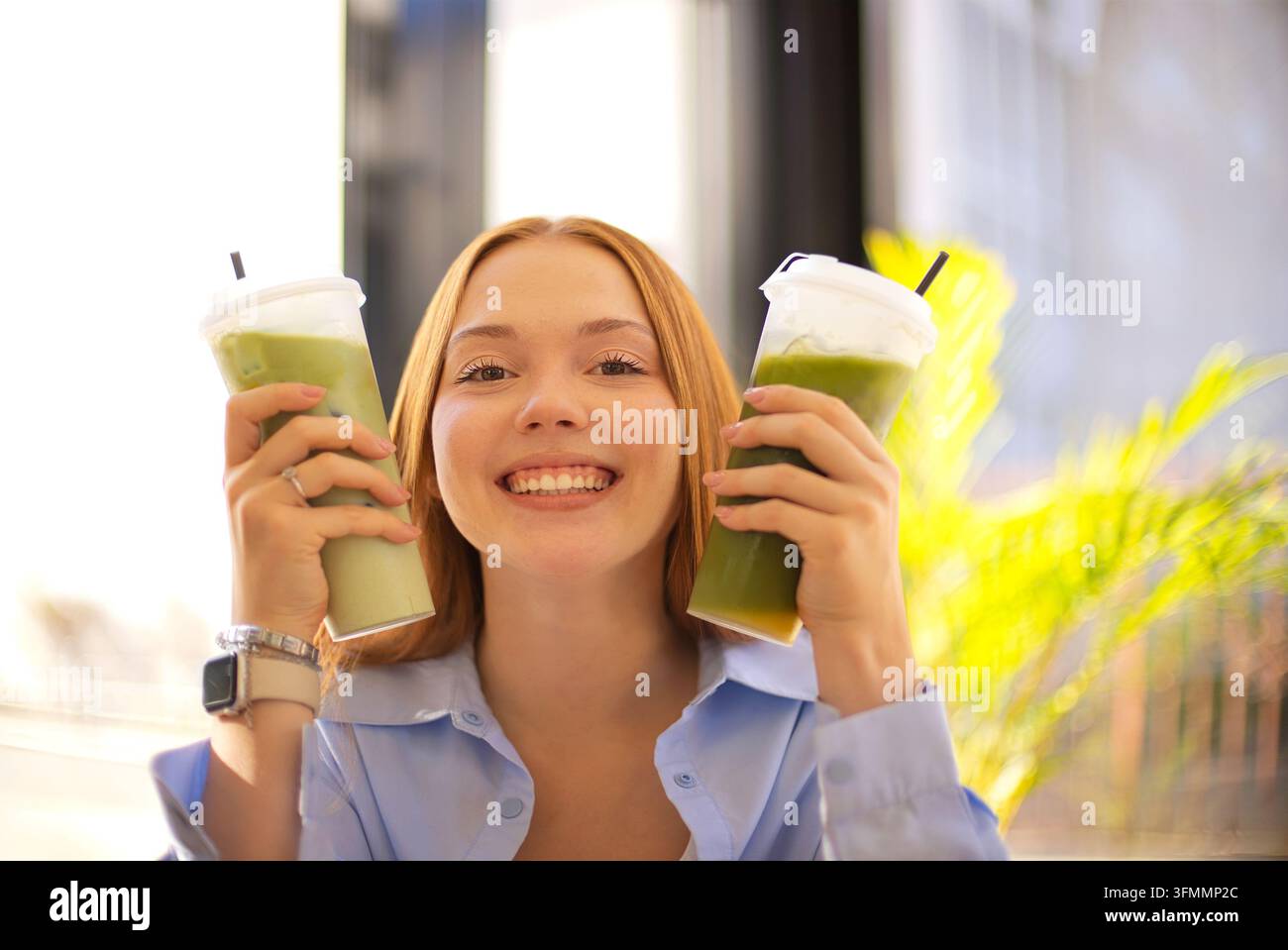 Femme souriante soulevant deux boissons mélangées vertes garnies de crème fouettée tout en se tenant devant des fenêtres lumineuses et un feuillage tropical ensoleillé à l'intérieur Banque D'Images
