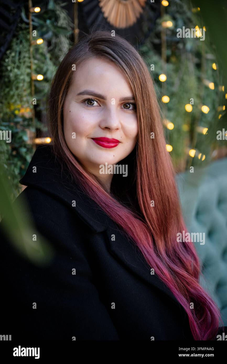 Portrait d'une femme souriante aux cheveux bruns longs et pointes roses, au rouge à lèvres rouge et au manteau noir, posée à côté d'un sapin de Noël décoré à la lueur chaude Banque D'Images