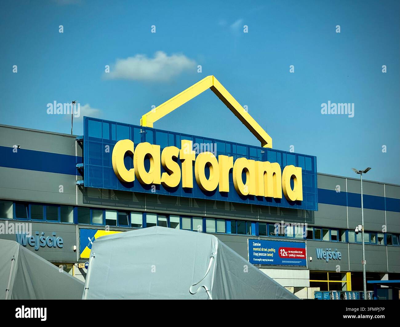 Prise de vue en contre-plongée d'un panneau du magasin de bricolage Castorama avec des lettres 3D jaunes sur un cadre métallique triangulaire bleu foncé, se détachant sur un ciel bleu - Image de stock capturée avec un smartphone