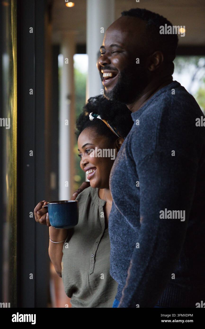 Couple africain debout près d'une grande porte vitrée à la maison, la femme tenant une tasse bleue et riant Banque D'Images