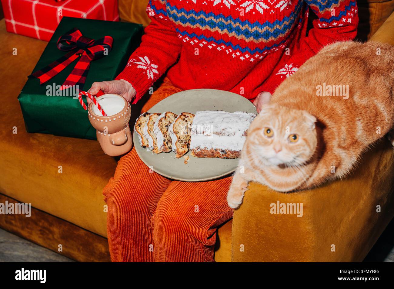 Femme en pantalon jaune vintage et pull festif assise sur un canapé marron avec un stollen de Noël traditionnel et du lait, tandis qu'un chat roux satisfait lou Banque D'Images