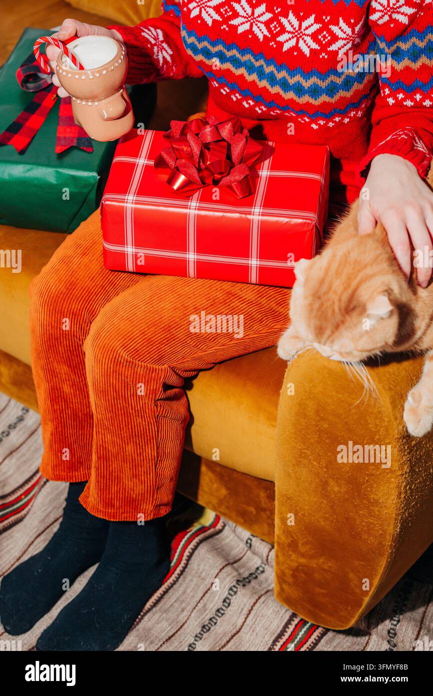 Scène de vacances chaleureuse d'une femme dans un pull festif et un pantalon en velours côtelé jaune tenant du lait à côté d'un chat gingembre, avec un cadeau de Noël emballé sur un b Banque D'Images