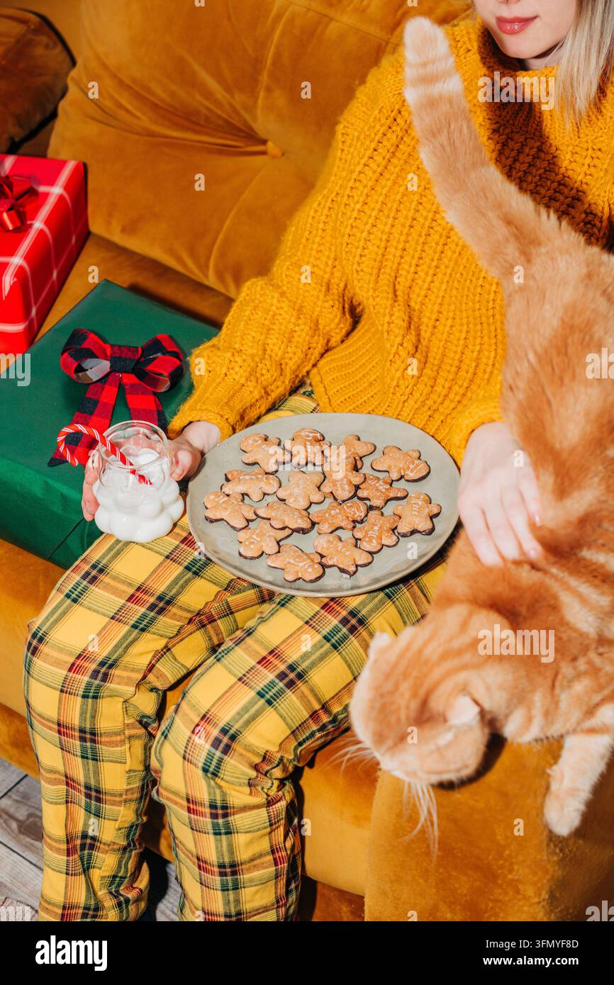Femme en pantalon à carreaux jaunes et un pull festif assise sur un canapé marron avec des biscuits sablés et du lait, tandis qu'un chat gingembre se prélasse à côté Banque D'Images