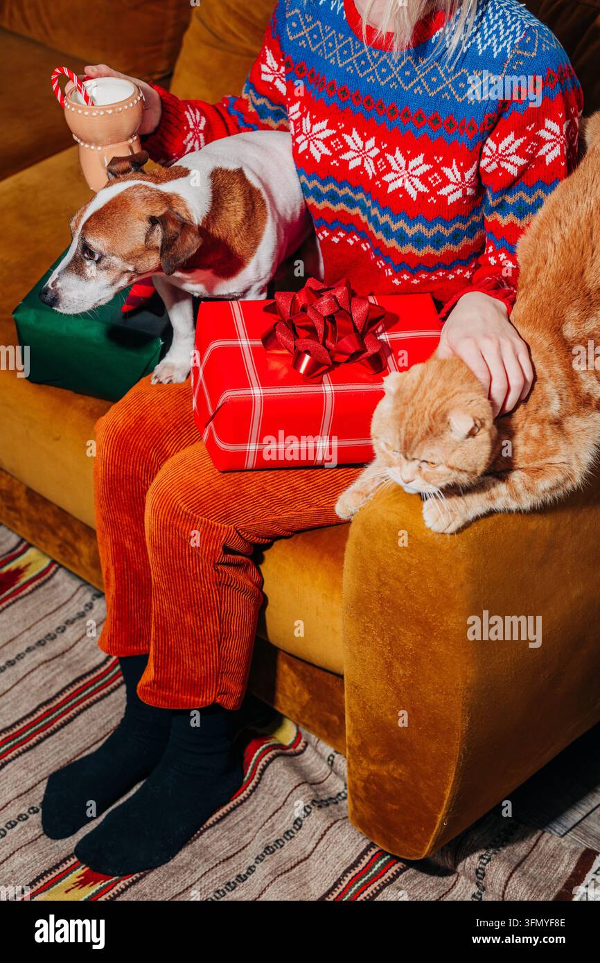 Femme en pull festif et pantalon vintage jaune assise sur un canapé marron avec un chat roux et un chien, tenant une boisson chaude à côté de cadeaux de Noël emballés Banque D'Images