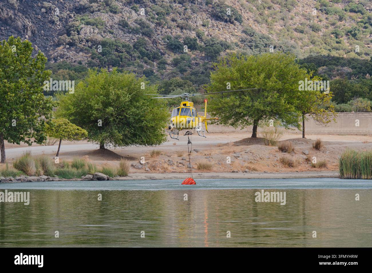 Hélicoptère de lutte contre les incendies jaune plane au-dessus d'un lac, abaissant un seau d'eau pour le remplir pendant les opérations de lutte contre les feux de forêt près de collines et d'arbres secs. Banque D'Images