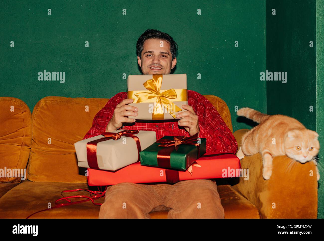 Homme souriant en chemise rouge assis sur un canapé orange tenant des cadeaux de Noël emballés tandis qu'un chat roux repose à ses côtés, créant une ambiance chaleureuse et festive à la maison Banque D'Images