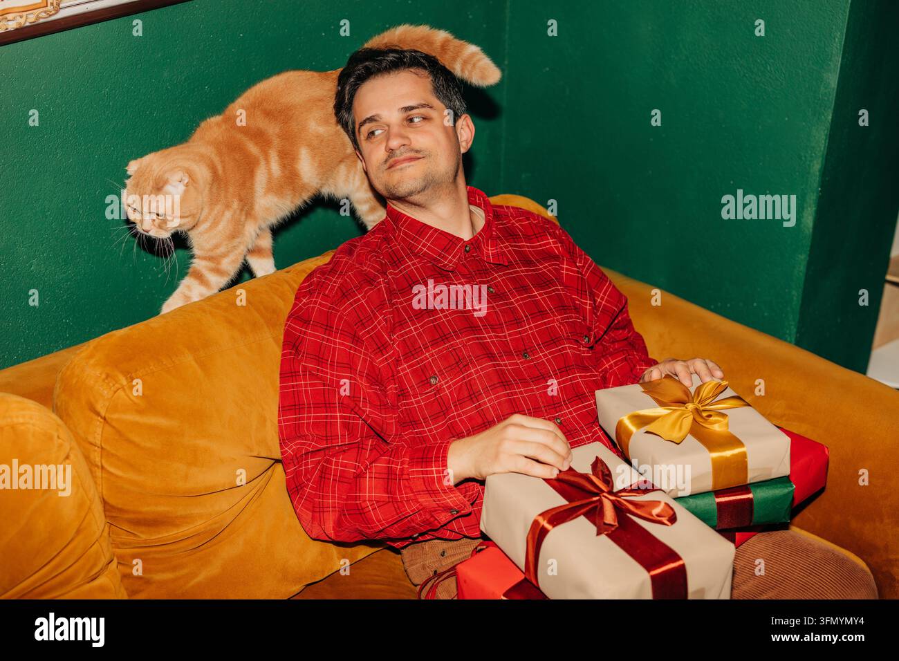 Jeune homme en chemise rouge assis sur un canapé moutarde tenant des cadeaux de Noël emballés tandis qu'un chat orange marche derrière lui dans un intérieur confortable. Banque D'Images